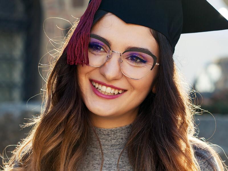Photo of a girl with glasses in a cap and gown smiling. 